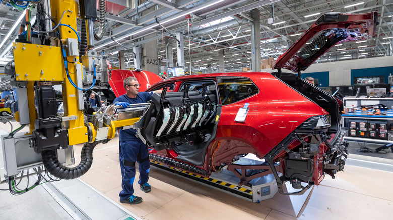 A photo of the rear three-quarters of a red iX3 being assembled with a robot arm handing a person the rear seats for installation