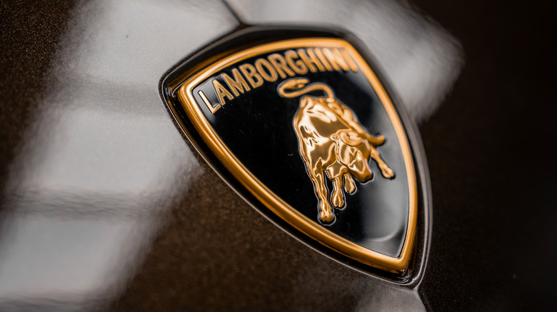 Riga, Latvia. April 15, 2025. A detailed view of the Lamborghini logo featuring a golden bull on a black shield with gold accents, set against a glossy dark car hood with light reflections.