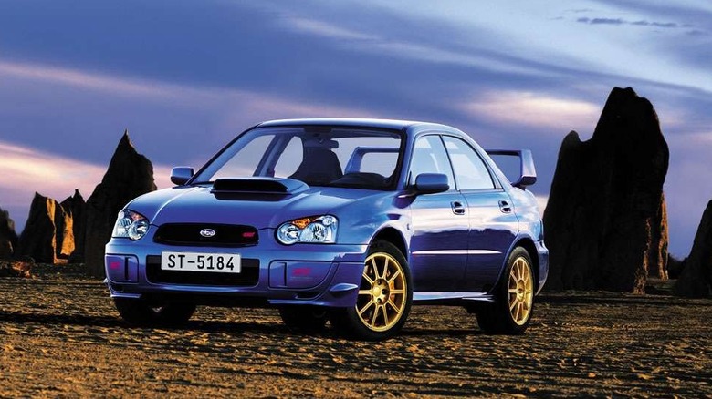 A front three-quarters shot of a blobeye 2004 Subaru WRX STIparked on dirt in front of vertical rocks and a cloudy sky at dusk