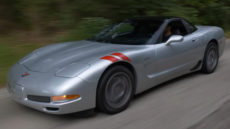 A silver C5 Corvette Z06 fixed-roof coupe with motion blur, featuring dual red hash marks on the front-left fender as it drives down a two-lane road.
