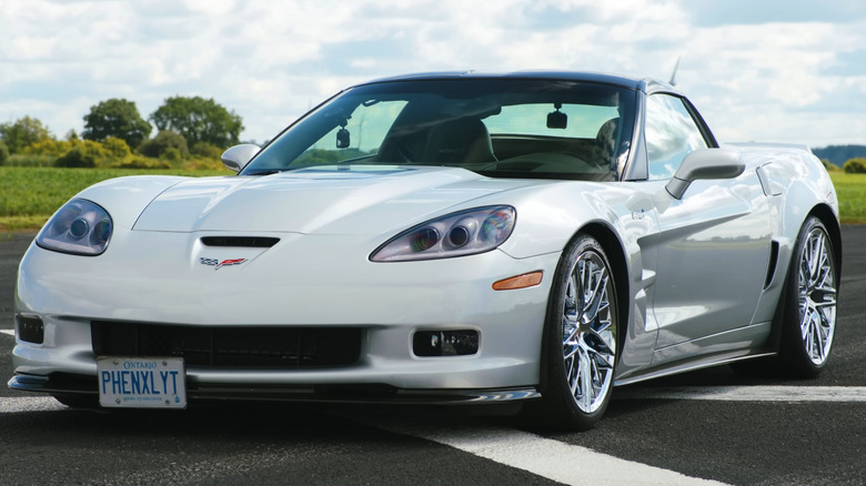 A silver C6 Corvette ZR1 with chrome multi-spoke wheels, parked on an asphalt track under a bright, cloudy sky.