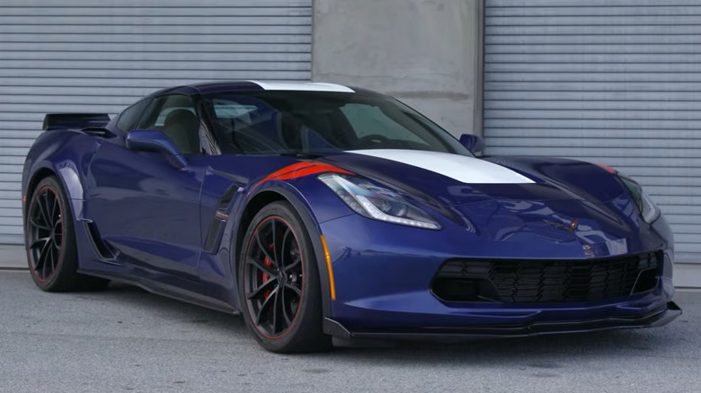 A dark blue C7 Corvette Grand Sport featuring a wide white center racing stripe and red fender hash marks, parked in front of closed metal garage doors.