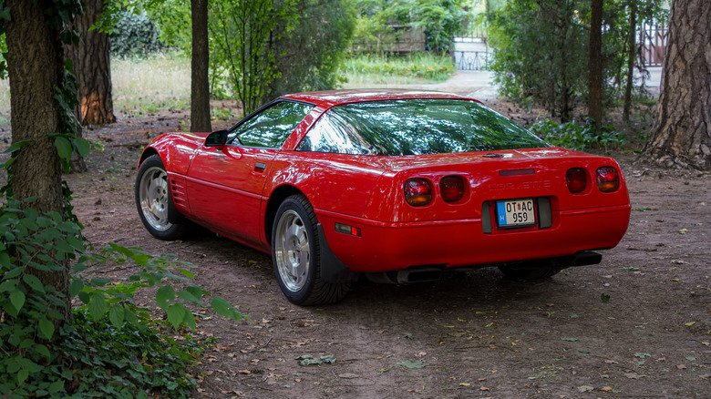 A red C4-generation Corvette seen from a rear three-quarter angle in a wooded setting