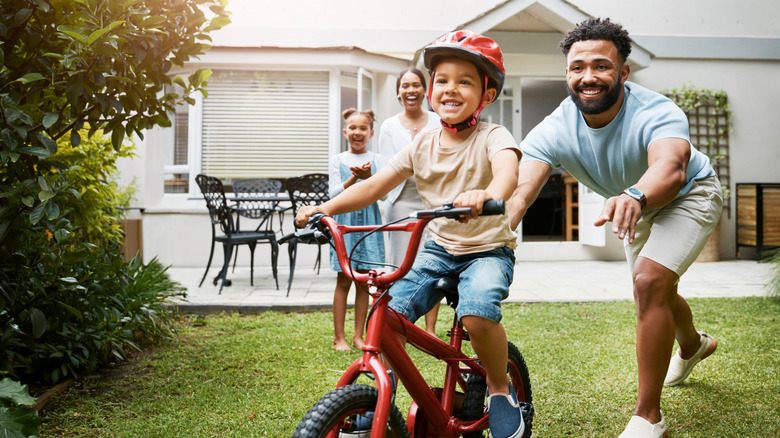 A proud dad teaches his young son to ride a bicycle outside their home while wearing a helmet for safety.