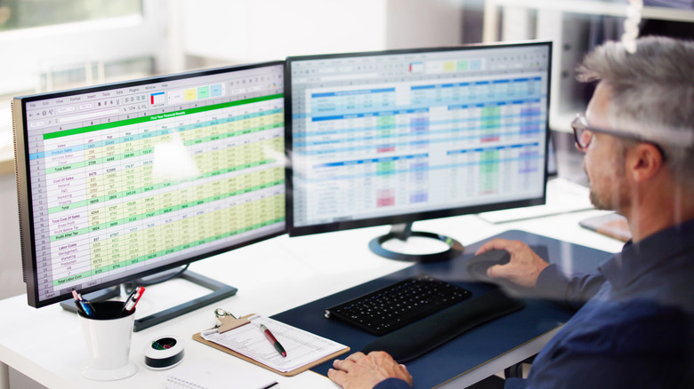 A man sitting at a desk with two computer screens showing data in a spreadsheet