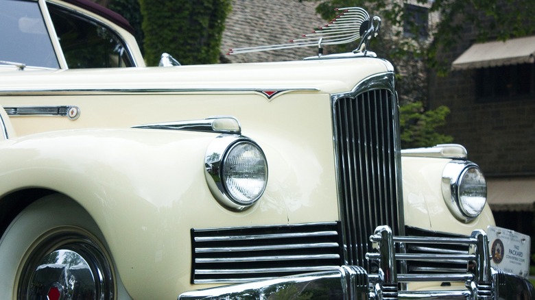 An ornate swan hood ornament graces the hood of a pale yellow 1942 Packard, featured at the Eyes on Design car show with the Edsel and Eleanor Ford mansion in the background.